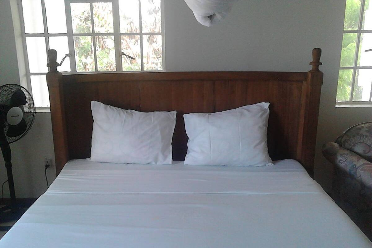 A bed with a wooden headboard and white bedding in a room with a window and a standing fan.