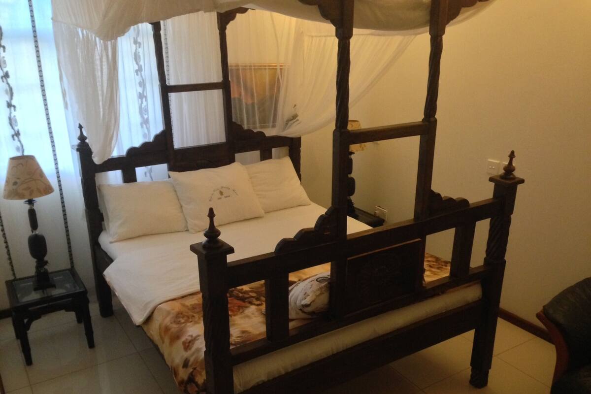 A four-poster bed with a canopy, a bedside table with a lamp, and a chair in a room with tiled flooring.
