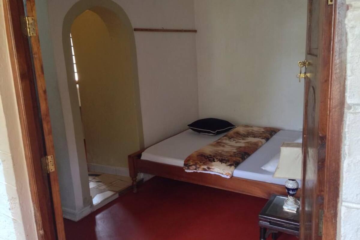A small, single-bed room with a wooden frame, a white mattress, a black pillow, a patterned blanket, a small bedside table with a lamp, and a door with a brass handle.