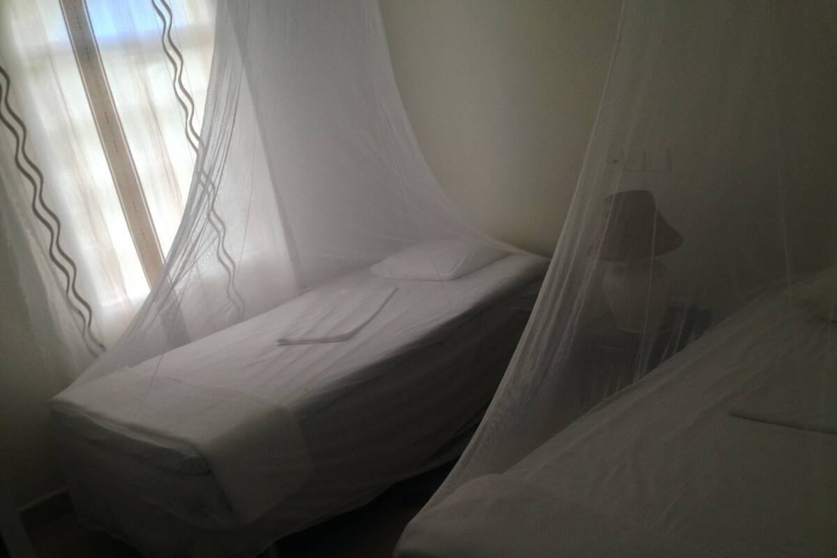 A dimly lit room with two single beds covered in white sheets, a window with sheer curtains, and a visible mosquito net.