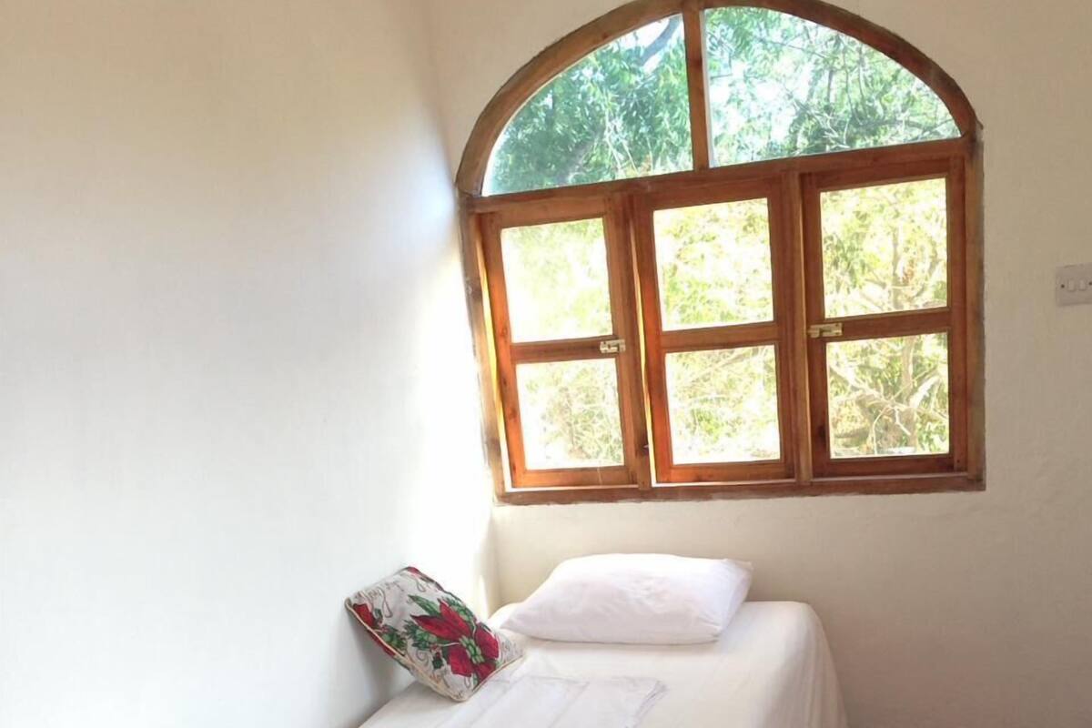 A single bed with a white bedspread and a floral pillow, a small table with a white cloth, and a window with a view of greenery.