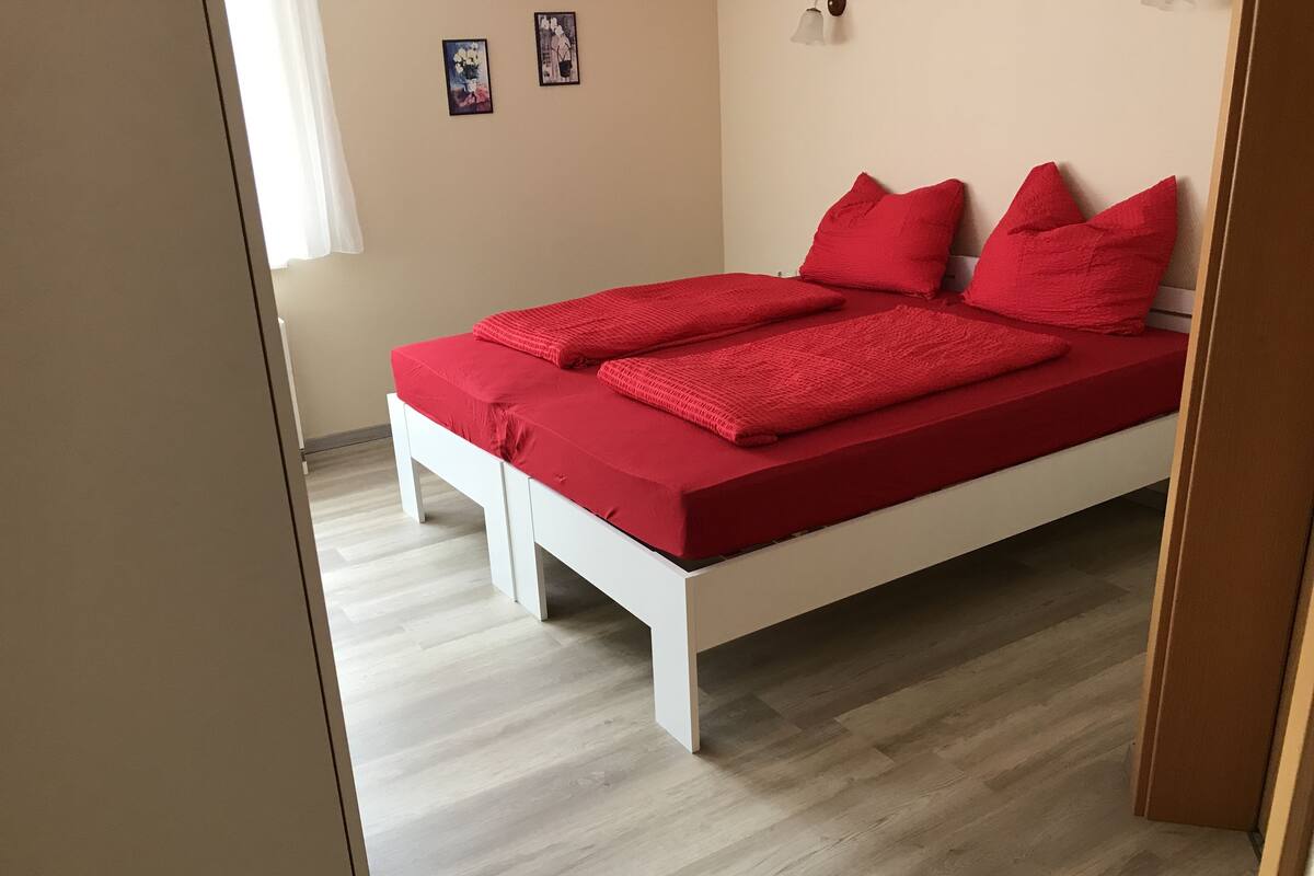 A bed with a red bedspread and pillows in a room with light wooden flooring and beige walls.