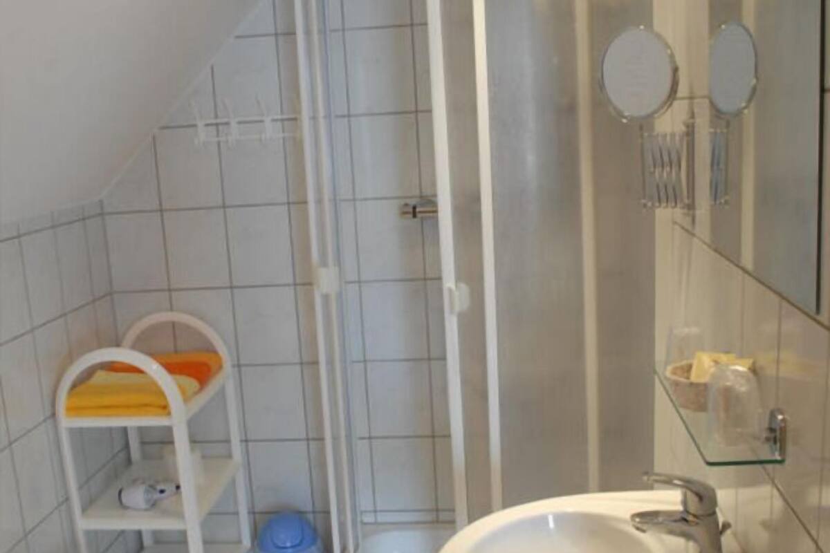 A bathroom with a white sink, a mirror, a shower, and a small white shelf with a yellow towel.