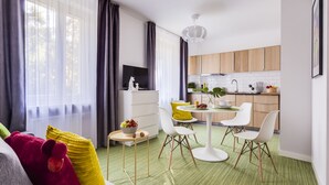 Una sala moderna con un sofá gris, cojines amarillos y rojos, una pequeña mesa redonda con frutas, y un comedor con sillas blancas y una alfombra verde.