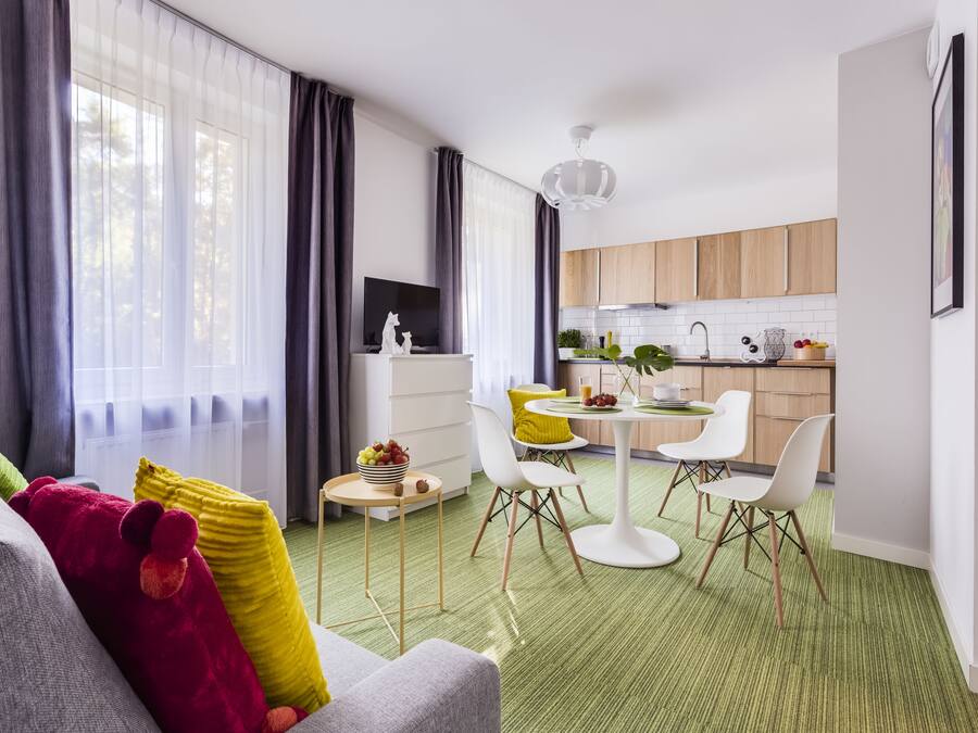 A modern living room with a grey sofa, yellow and red cushions, a small round table with fruits, and a dining area with white chairs and a green carpet.