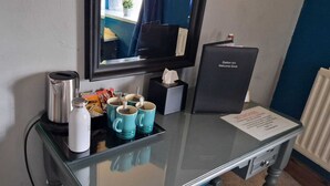 A hotel room with a glass table displaying a coffee maker, a water bottle, and several mugs. A welcome book and a no-smoking sign are also visible.