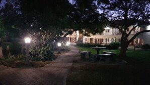 Un sendero pavimentado con bancos y un jardín bien cuidado, que conduce a un edificio con varias ventanas y una escalera.