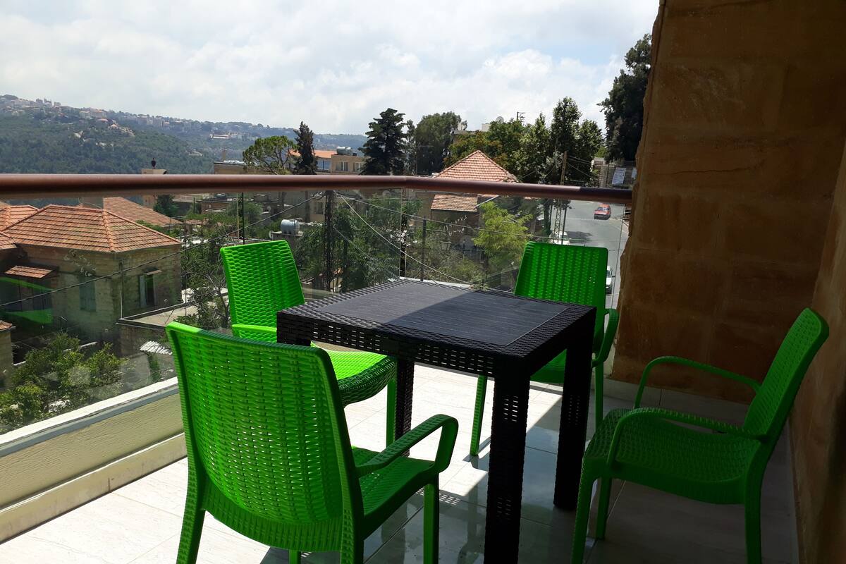 A balcony with a table and chairs, overlooking a cityscape with buildings and trees.