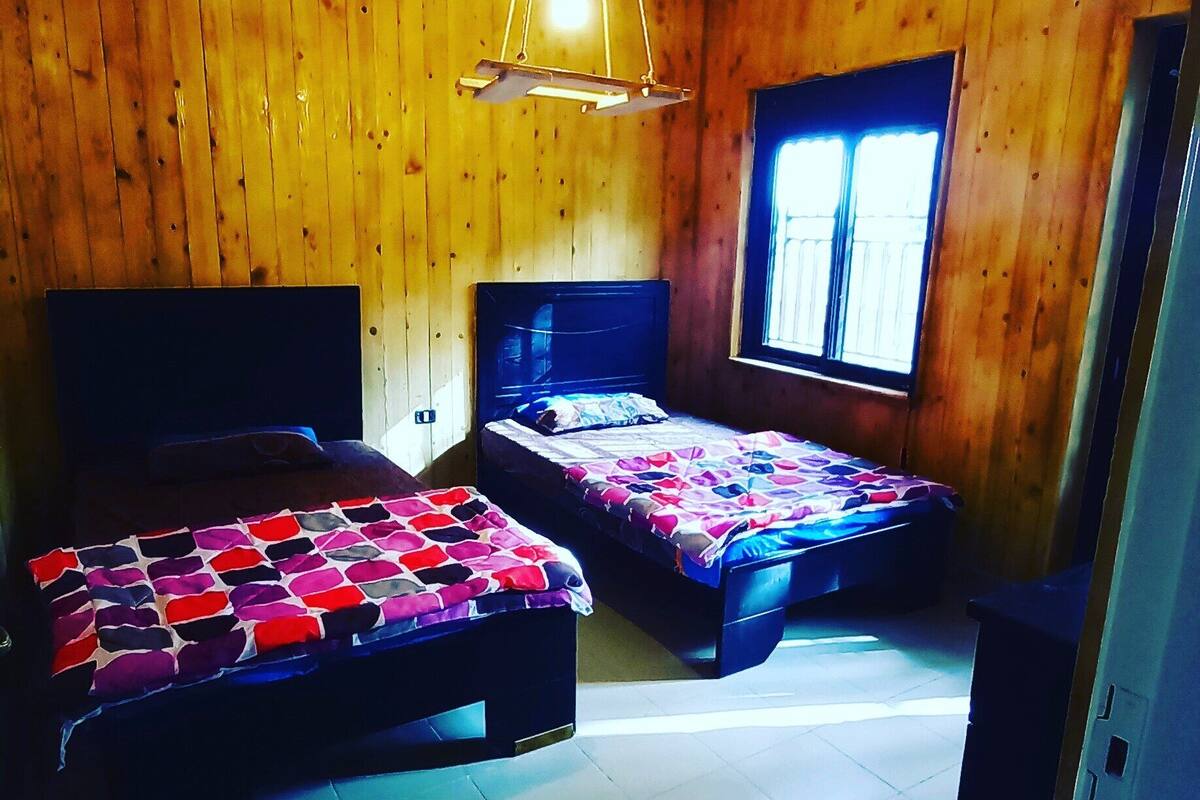 A room with two beds, wooden walls, a window, and a hanging light fixture.