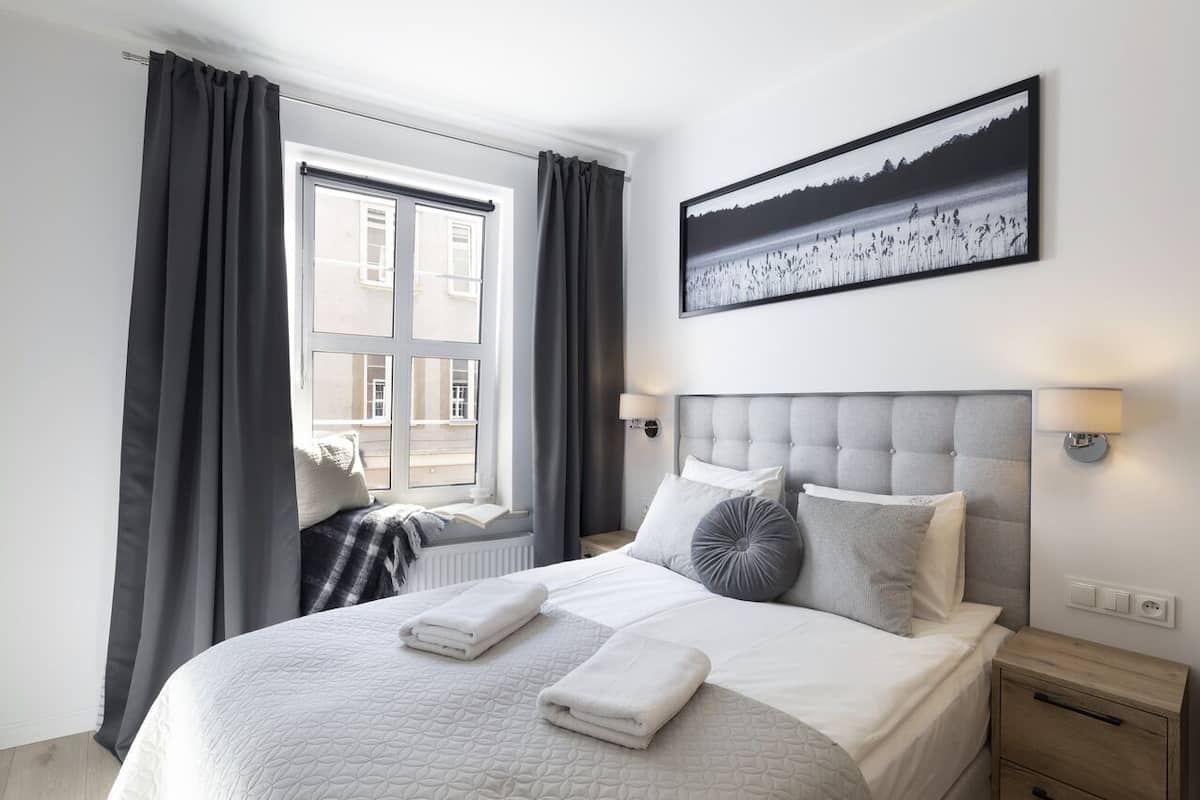 Una cama bien hecha con ropa blanca y dos toallas dobladas, cabecera gris, lámpara de noche y ventana con cortinas grises.