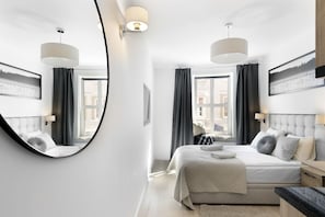 A modern bedroom with a large round mirror, two beds with matching headboards, a large window with curtains, and wall-mounted lighting.