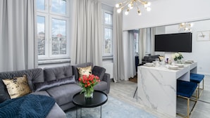 Ein modernes Wohnzimmer mit einem grauen Sofa, einem schwarzen Couchtisch und einer Vase mit roten Blumen. Es gibt einen Essbereich mit einer weißen Marmorinsel und blauen Stühlen.