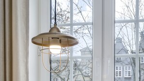 Ein gemütliches Zimmer mit großem Fenster, bequemen Sessel, Tisch mit Buch und Tasse sowie einer Pendelleuchte.