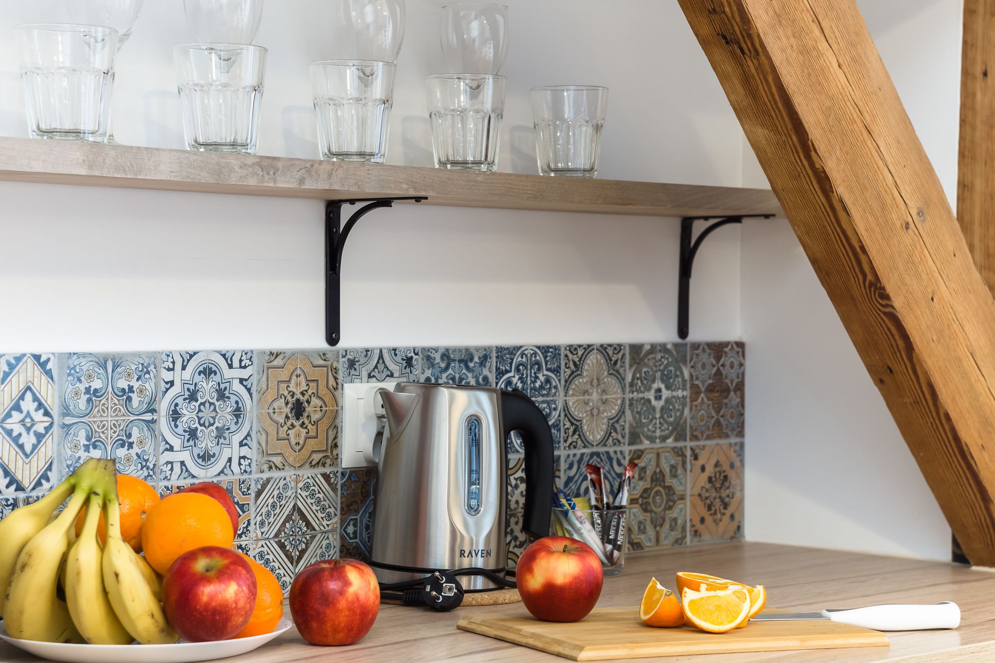 Una cocina con salpicadero de azulejos, una tetera, una tabla de cortar con fruta y un estante de madera con vasos.