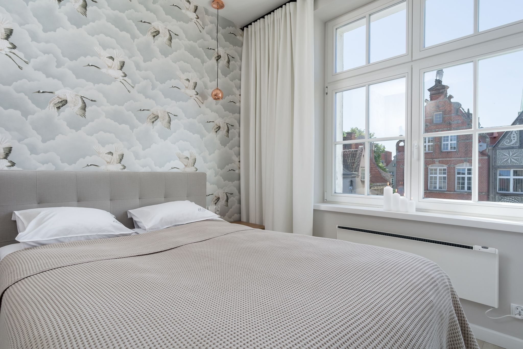 A bedroom with a large bed, a window with a view of buildings, and a patterned wall.