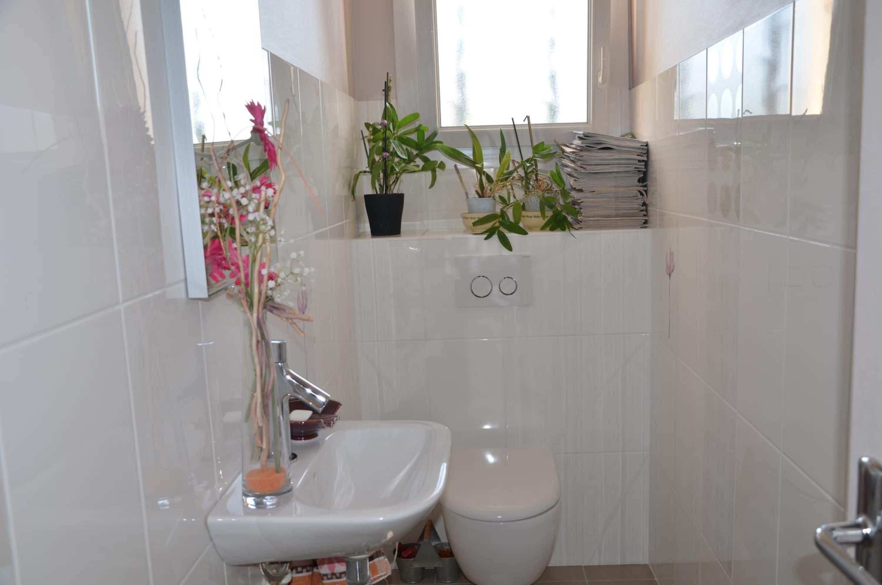 Een badkamer met een wit wastafel, toilet en een spiegel boven het wastafel. Er staan planten op de vensterbank en een stapel papier aan de muur.