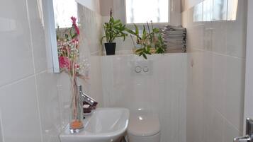 Een badkamer met een wit wastafel, toilet en een spiegel boven het wastafel. Er staan planten op de vensterbank en een stapel papier aan de muur.