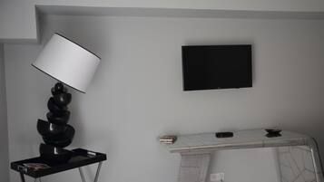 Una habitación moderna con un televisor de pantalla plana montado en la pared, una lámpara negra de diseño sobre un soporte y un elegante escritorio blanco con estampado de mármol.