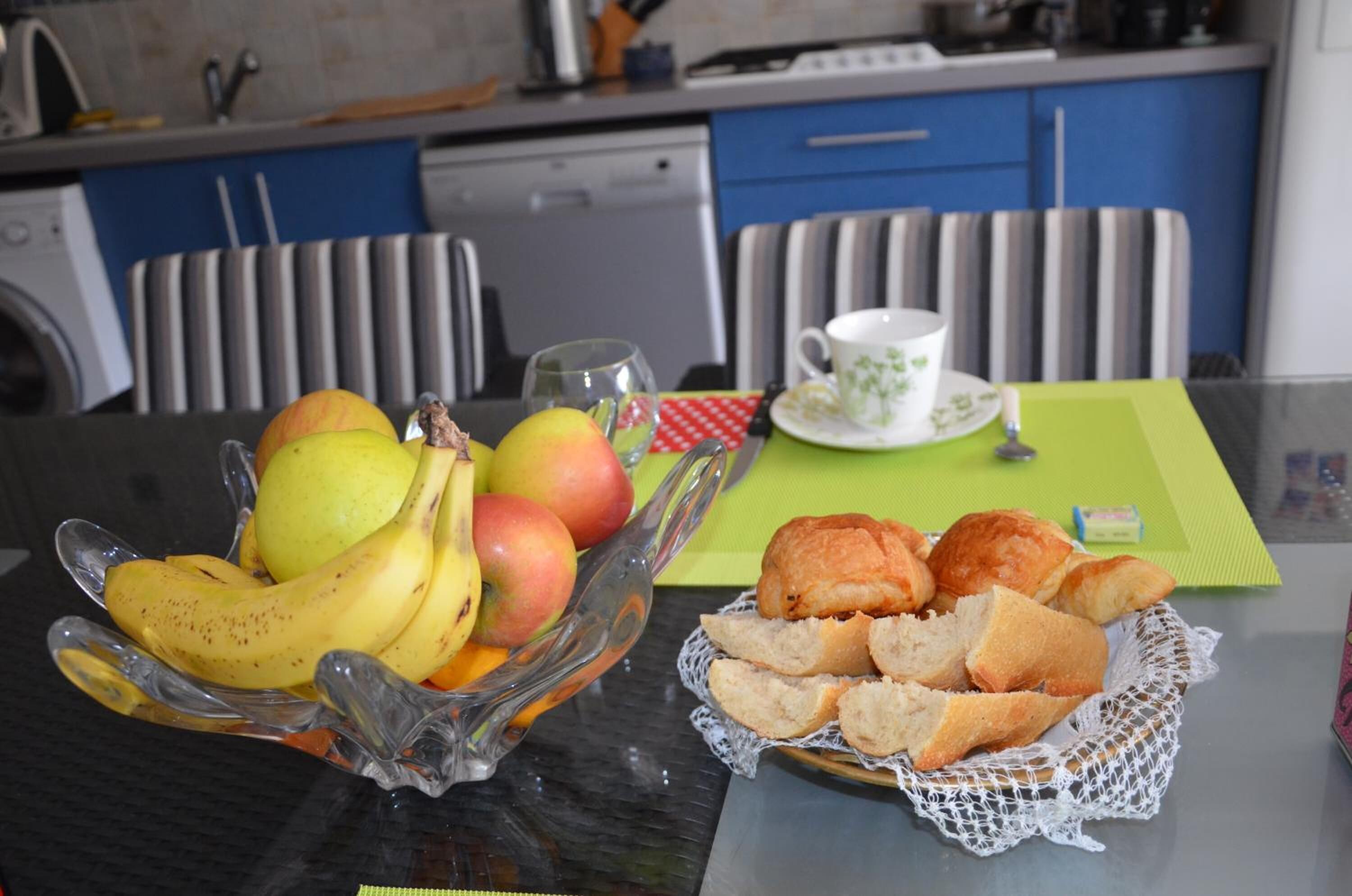 Una mesa de comedor con un frutero que contiene plátanos y manzanas, una cesta de panecillos y una taza de té sobre un mantel individual verde.