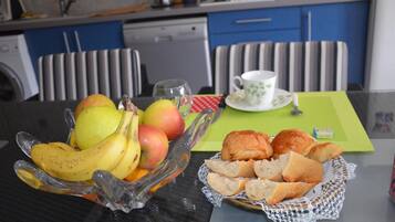 Una mesa de comedor con un frutero que contiene plátanos y manzanas, una cesta de panecillos y una taza de té sobre un mantel individual verde.