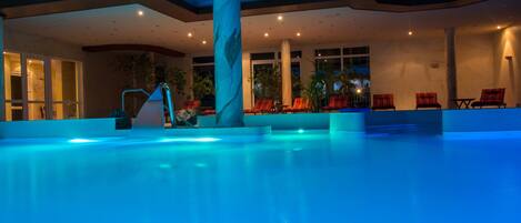 Una piscina interior con un columnado de mármol, sillas reclinables rojas e iluminación empotrada en el techo.