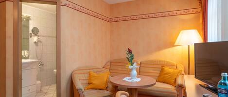 Habitación de hotel con ducha, sofá con cojines amarillos, una mesita redonda con un florero y un televisor sobre un mueble de madera.