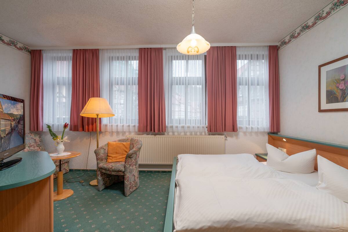 A hotel room with a bed, a chair, a desk, a lamp, and a window with curtains.