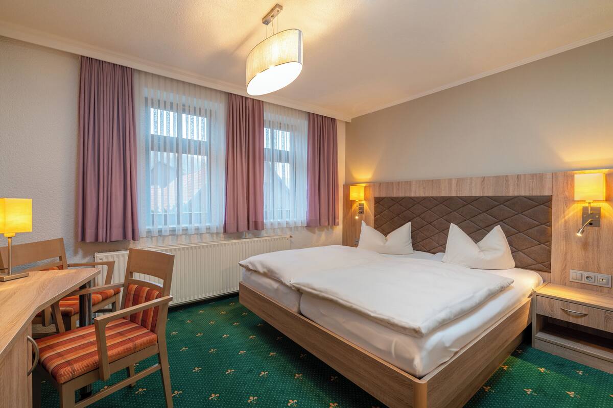 A hotel room with a bed, a desk with a chair, a lamp, and a window with curtains.