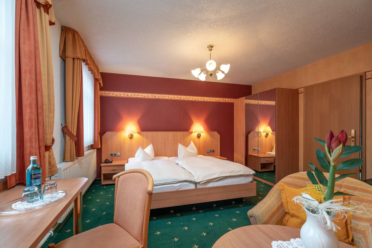 A hotel room with a bed, bedside tables, a desk, a chair, and a vase with flowers.