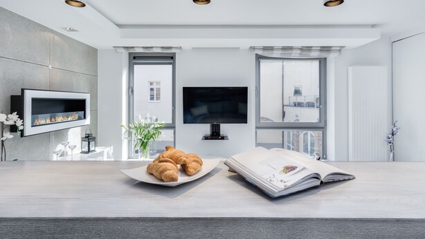 A modern room with a flat-screen TV, a fireplace, a large window, and a dining area with a plate of croissants and an open book. - Elite Apartments Old Town Prestige (Gdansk)