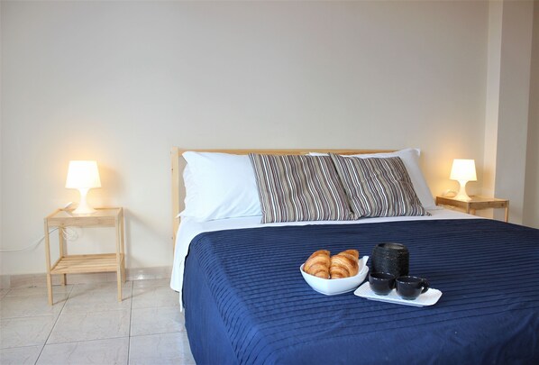 Una cama bien hecha con una colcha azul, dos lámparas de noche, un almohadón a rayas y una bandeja con croissants y tazas sobre la cama.
