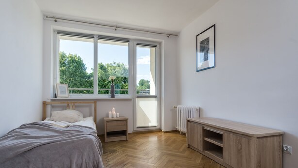 Un dormitorio con cama, mesita de noche, radiador y ventana con vistas a árboles.