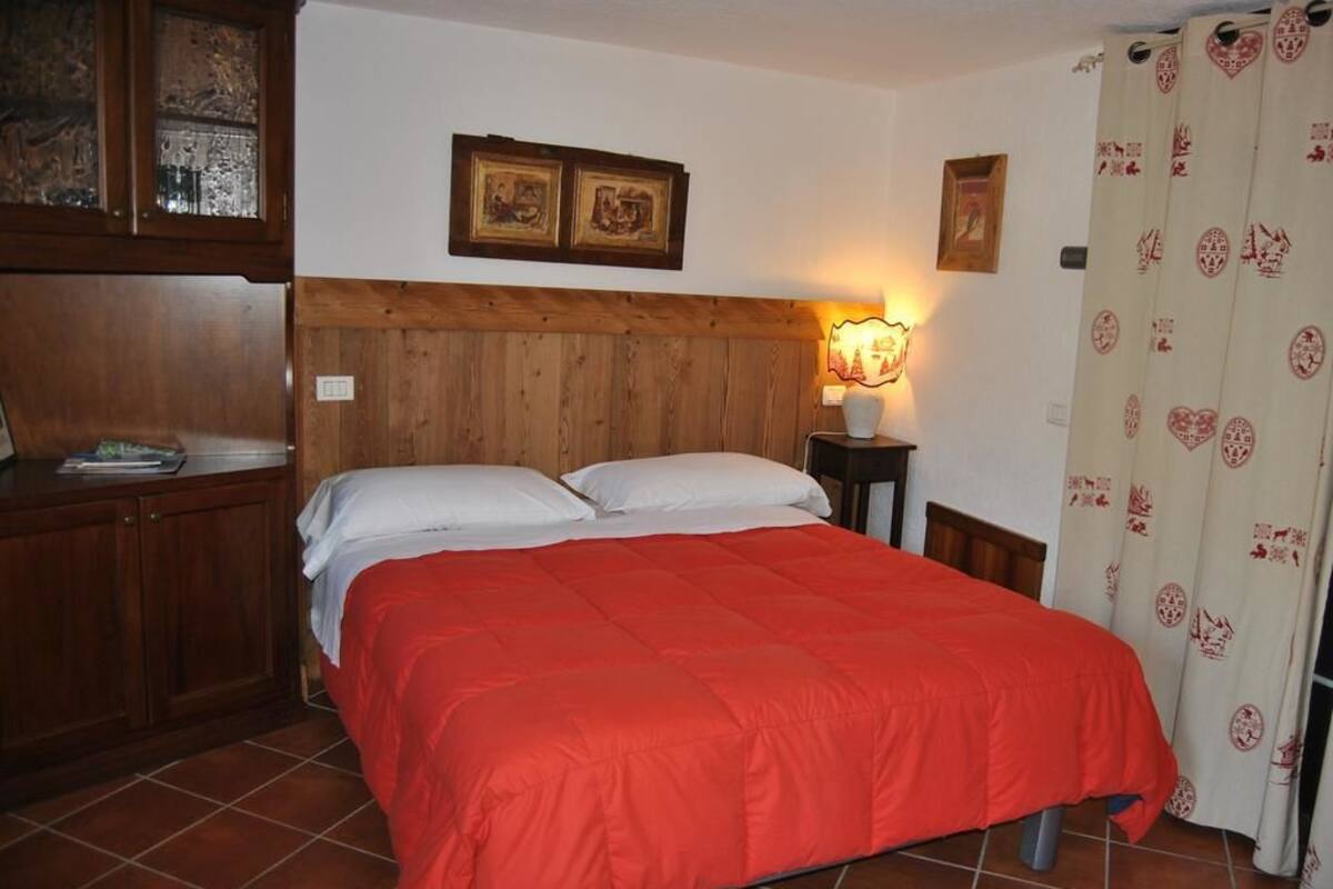 A bedroom with a wooden headboard, a bed with a red blanket, a nightstand with a lamp, and a curtain with a pattern.