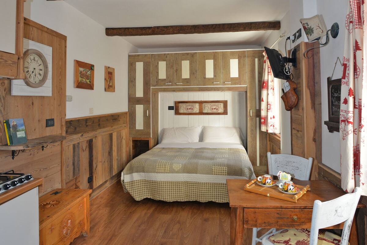 A rustic bedroom with a wooden bed, a wooden dresser, a small table with a chair, and a wall clock.
