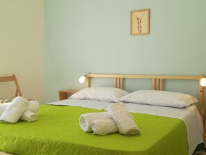 A neatly made bed with a green bedspread, two white pillows, and two rolled white towels. There is a wooden headboard, a bedside table with a lamp, and a framed picture on the wall.