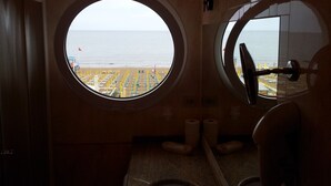Une pièce dotée d’une fenêtre circulaire donnant sur une plage avec des parasols et un poste de surveillance pour les sauveteurs.