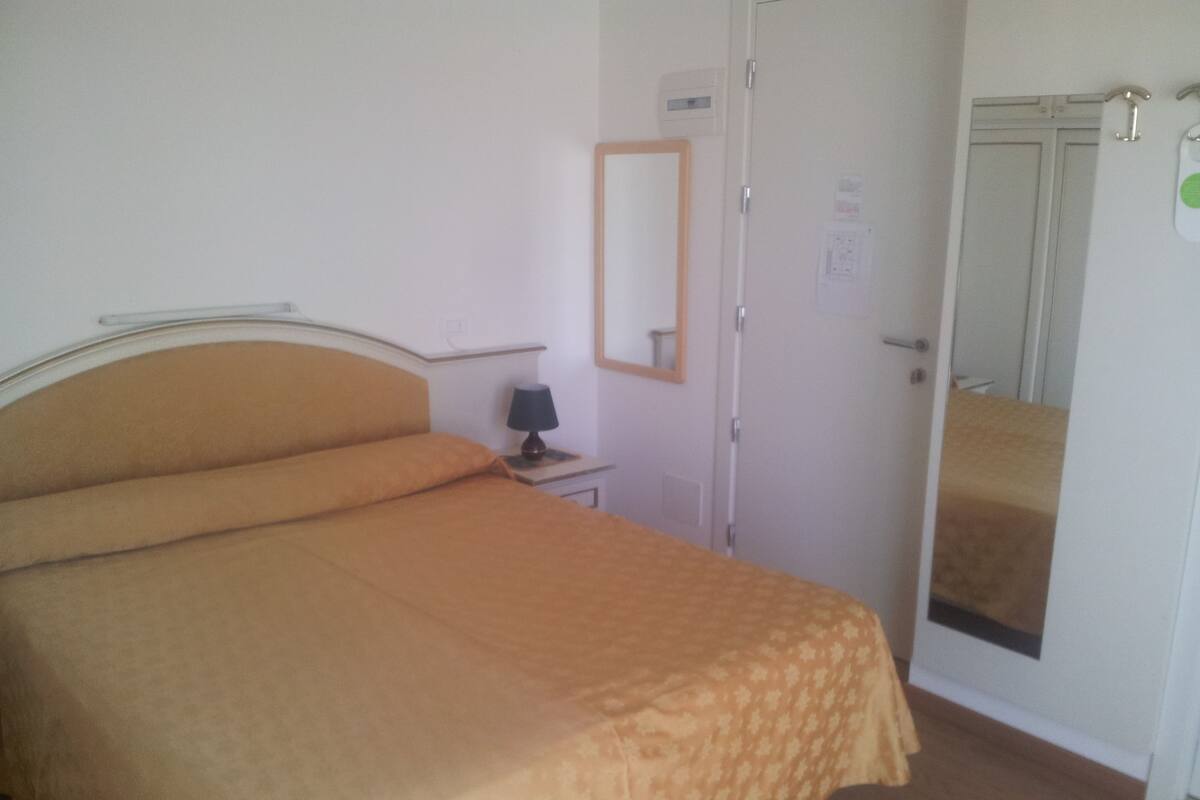 A bedroom with a bed, a bedside table, a lamp, a mirror, and a wardrobe.
