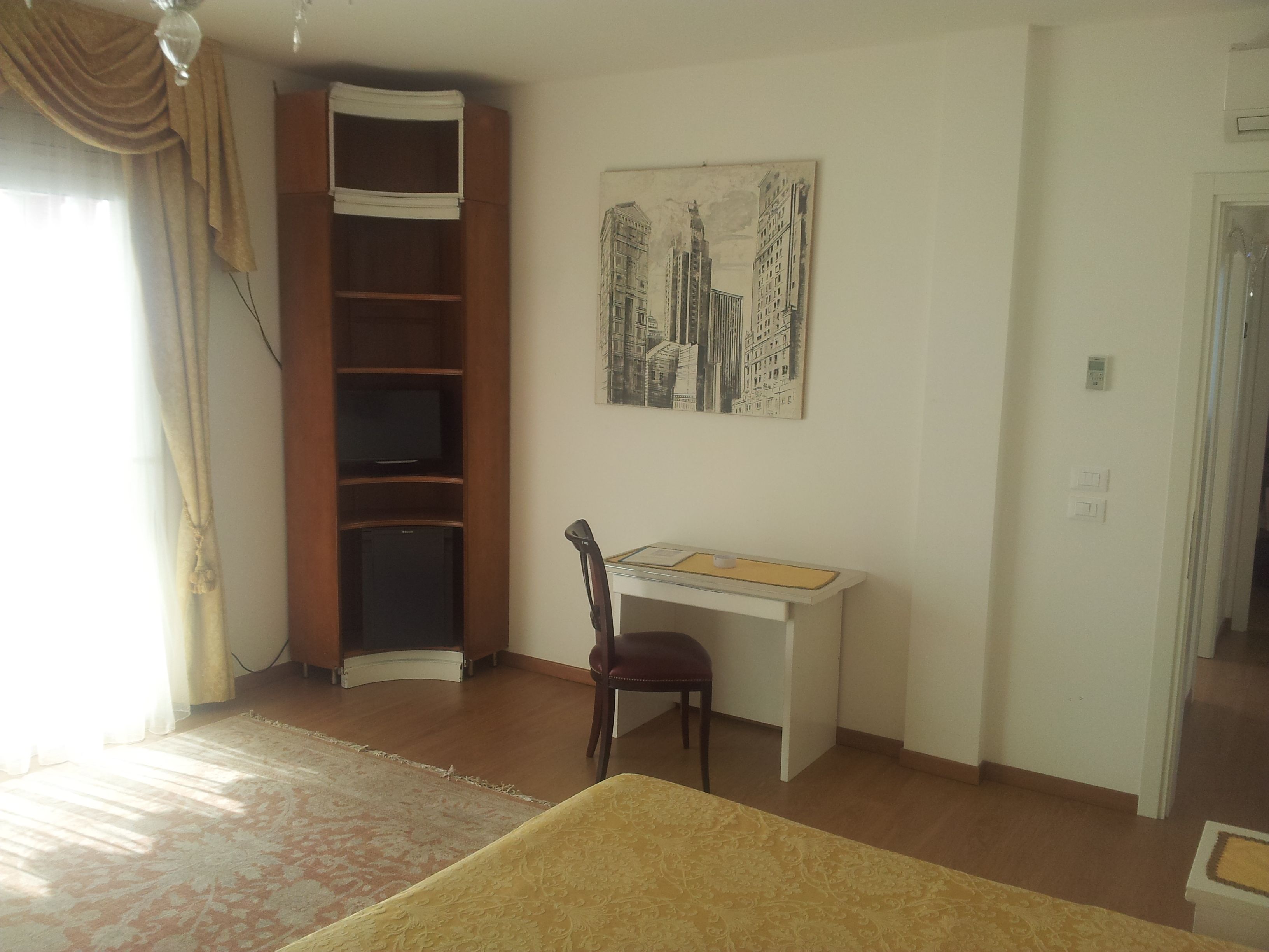 A room with a desk, chair, and a bed. There is a painting on the wall and a window with curtains.