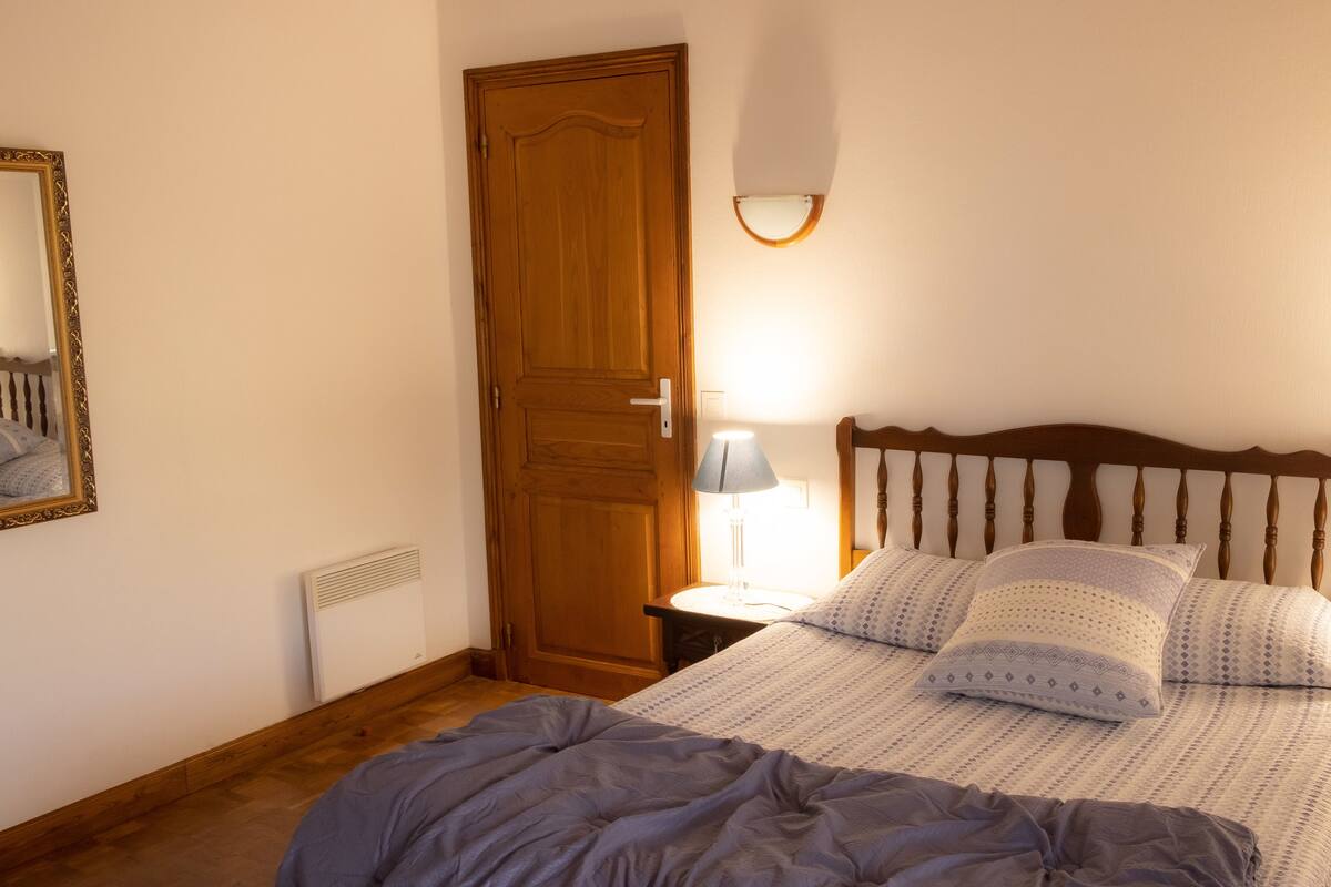 A bedroom with a wooden door, a bed with patterned bedding, two bedside lamps, and a mirror on the wall.