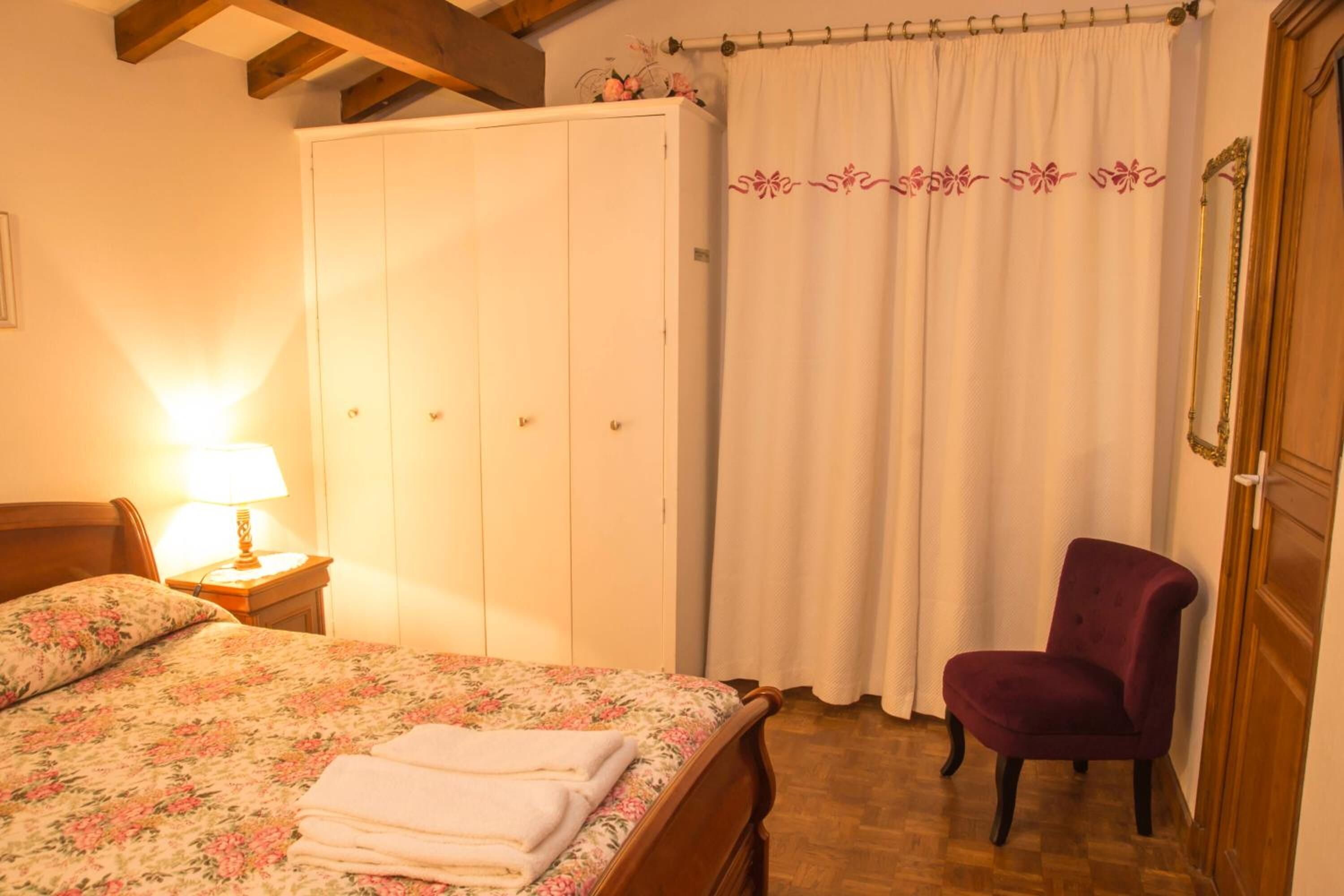Un dormitorio con cama, mesita de noche, armario, silla y cortina.