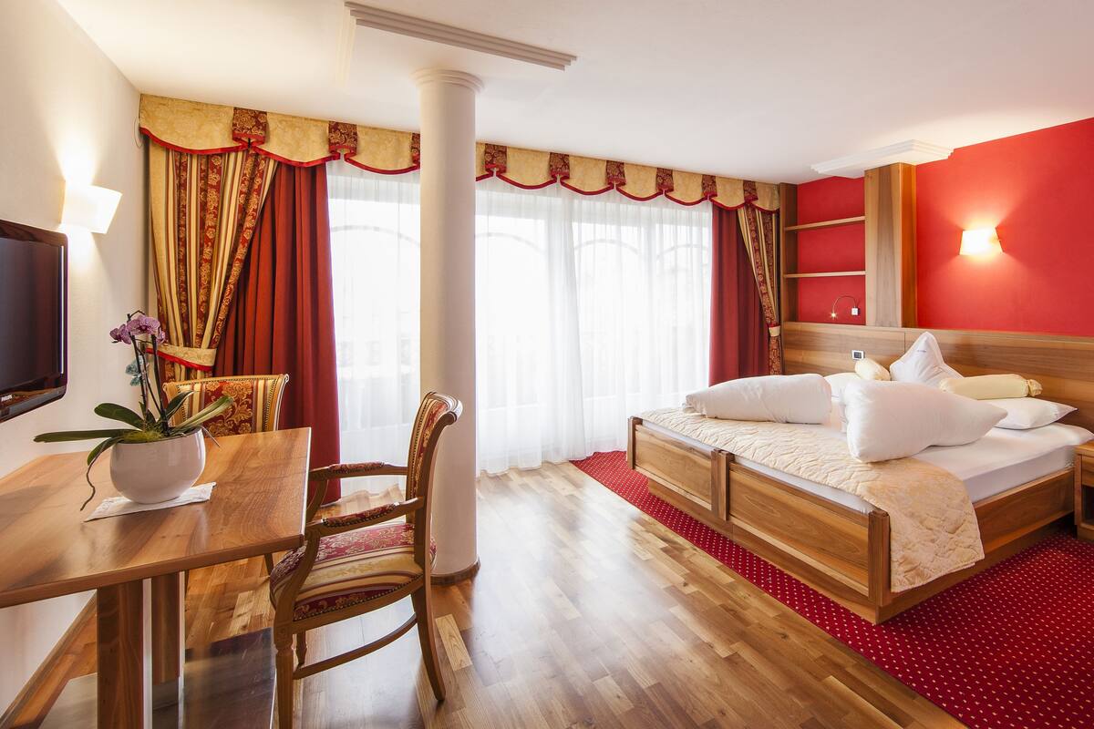 A hotel room with a wooden bed, a desk with a chair, a TV, and a red carpet.
