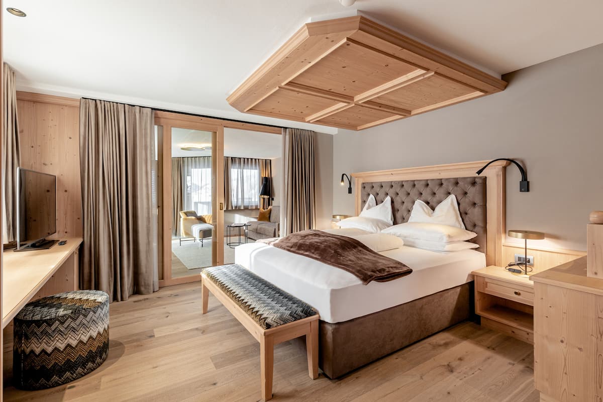 A modern hotel room with a large bed, wooden headboard, a desk with a chair, a bench, and a view of the outside through a sliding door.