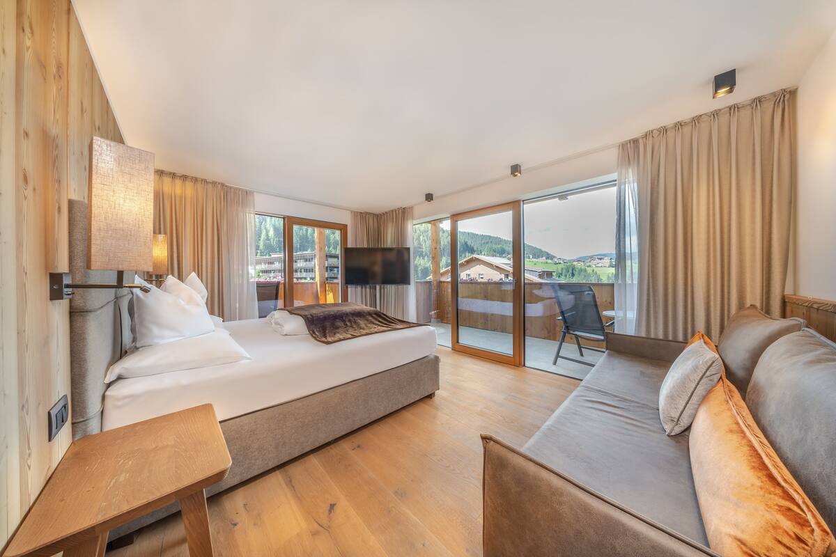 A modern hotel room with a large bed, a sofa, a small table, and a TV. The room has large windows with a view of the mountains.