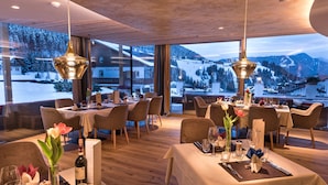 Ein Restaurant mit gedeckten Tischen, großen Fenstern mit Blick auf eine verschneite Berglandschaft und hängenden Pendelleuchten.