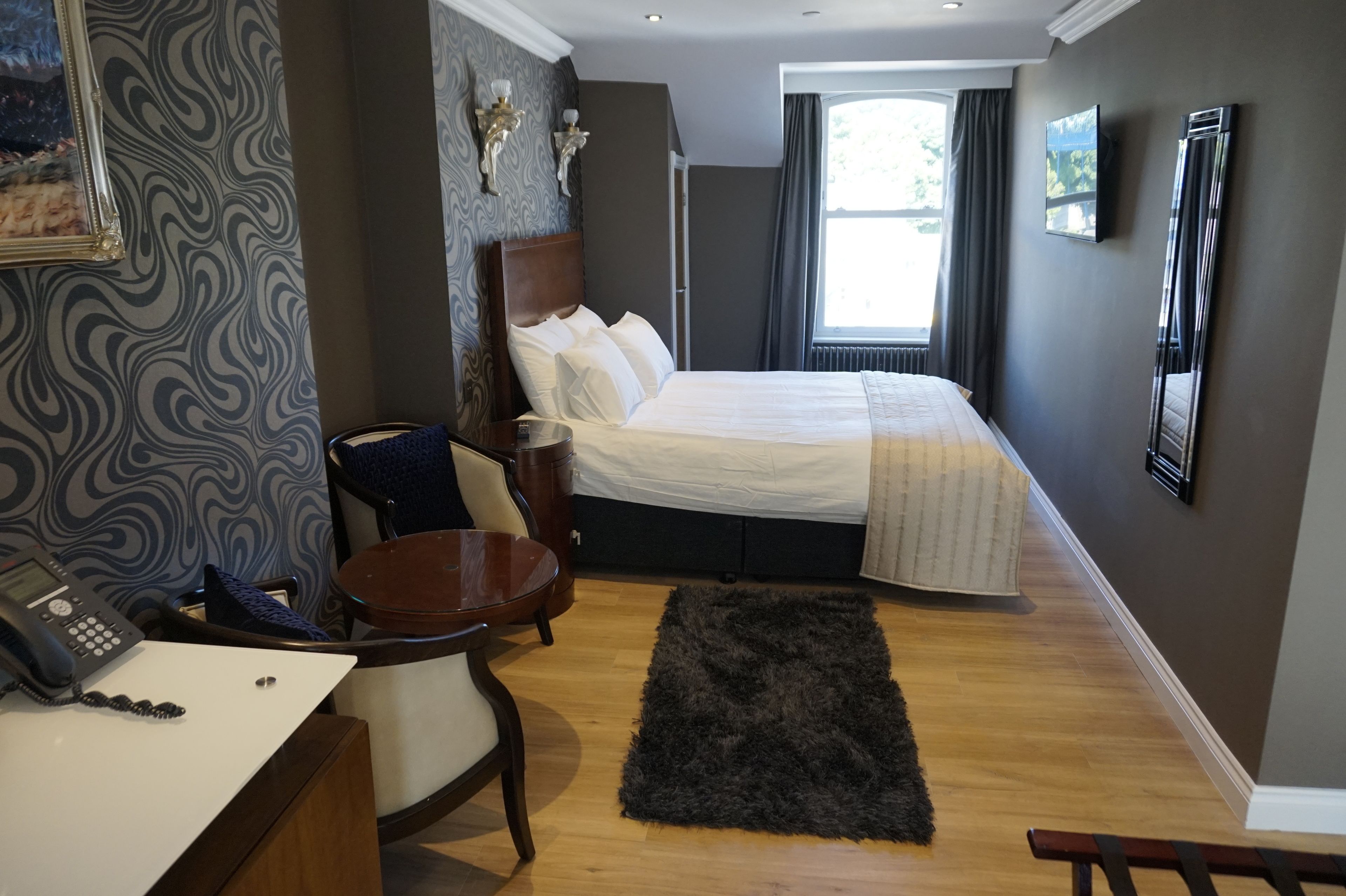 Ein Hotelzimmer mit einem großen Bett, einem Schreibtisch mit Telefon, einem Sessel, einem kleinen Tisch und einem dunklen Teppich auf dem Boden.