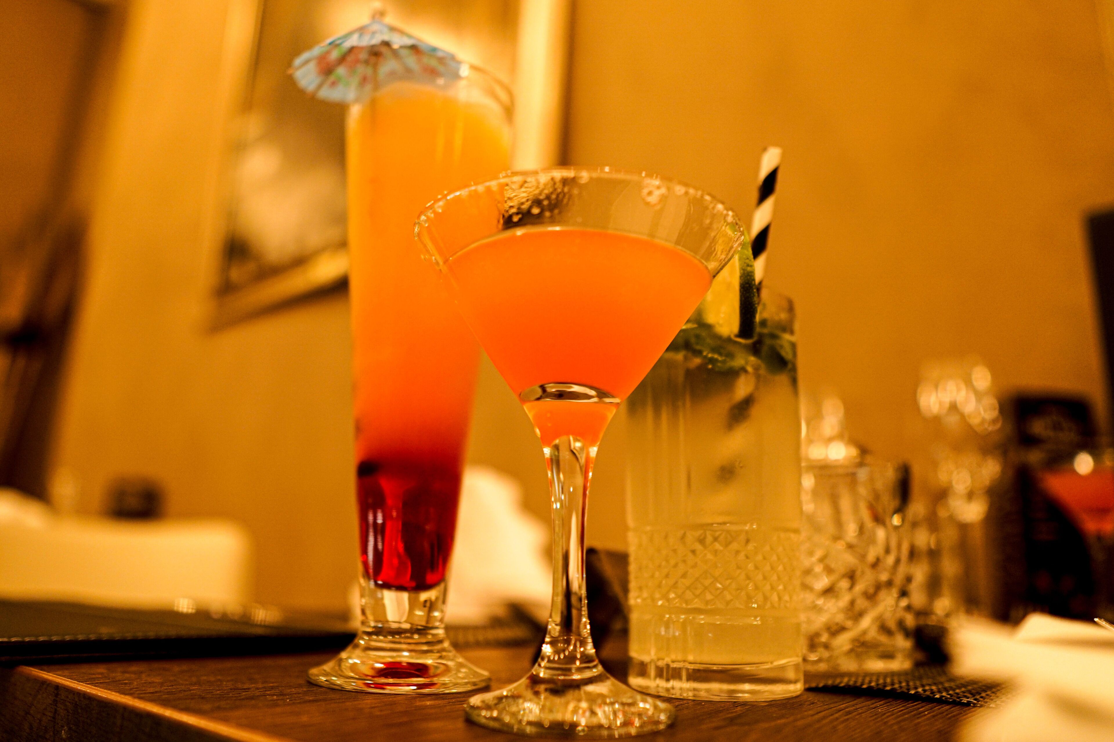 Una zona de bar con un vaso alto de bebida de naranja, una copa de martini con pajita y una jarra de agua.