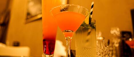 Una zona de bar con un vaso alto de bebida de naranja, una copa de martini con pajita y una jarra de agua.