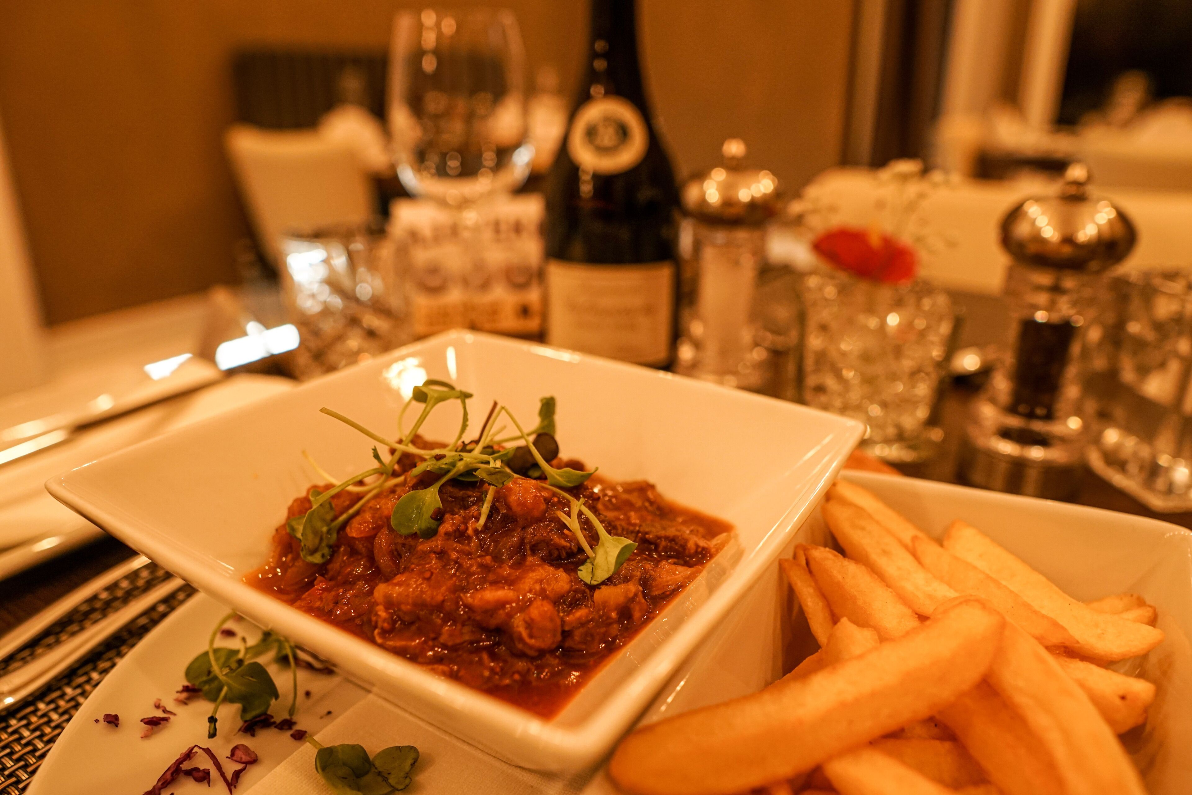 A dining table with a square plate of stew garnished with greens, a bowl of fries, and a bottle of wine.
