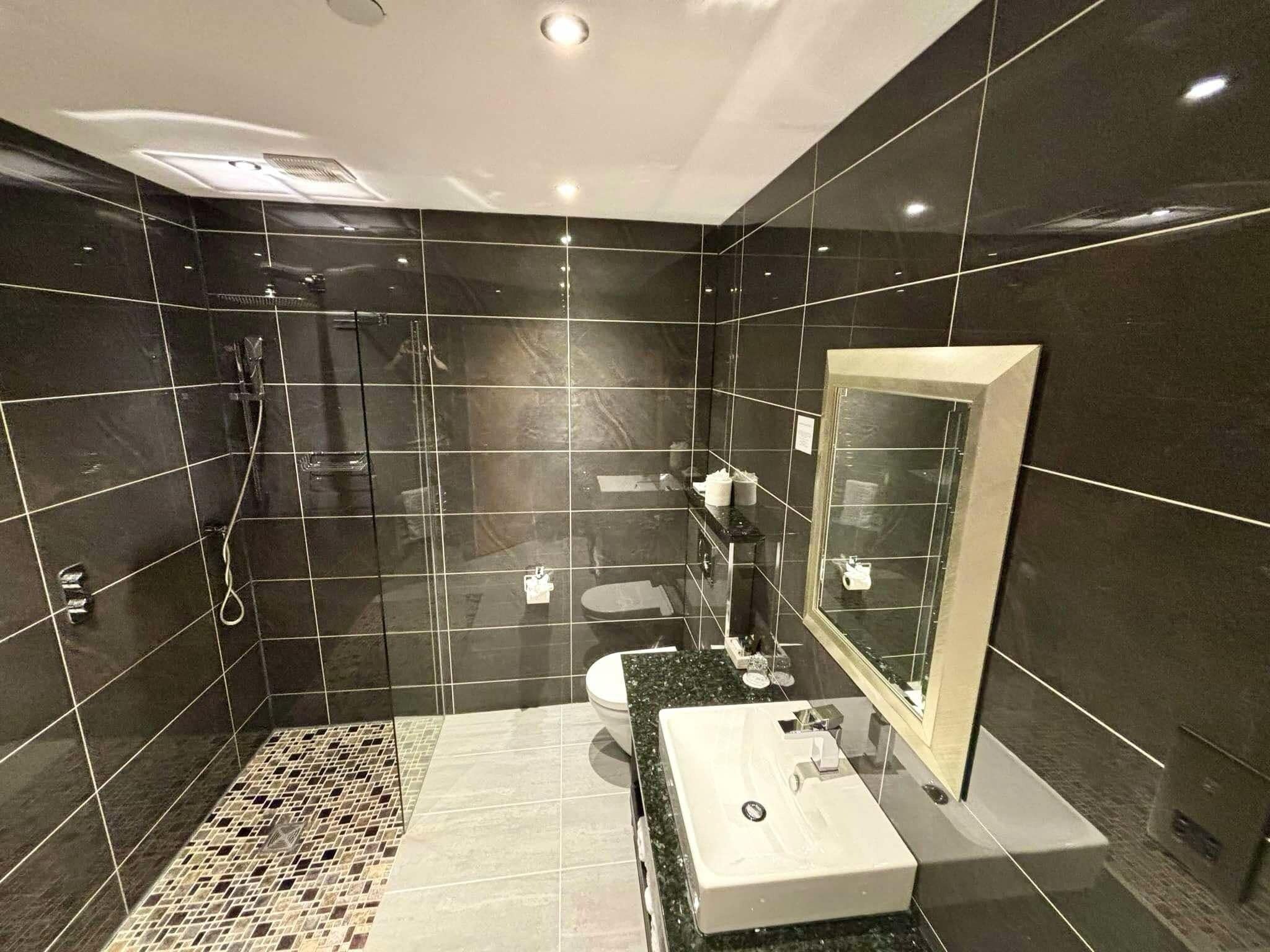 Un baño moderno con baldosas oscuras, una cabina de ducha de vidrio, un lavamanos con espejo y un suelo estampado.