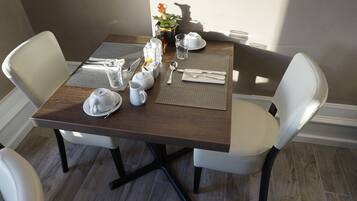 A dining table set with white plates, silverware, and a pitcher, surrounded by white chairs in a room with a wooden floor and a potted plant.